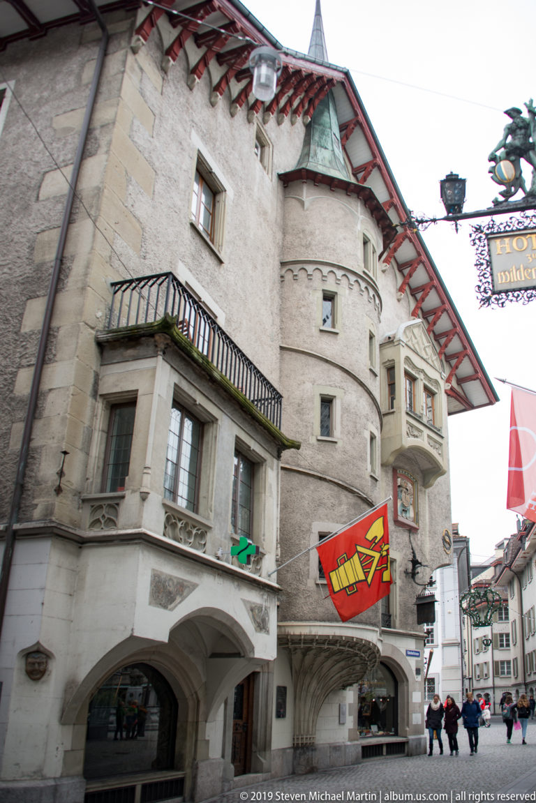 Close view of a historic Lucerne corner building with balcony, deep eaves, stone windows, and arched ground-floor arcade.