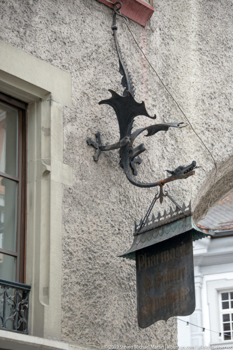 Black metal dragon-shaped pharmacy sign projecting from a rough plaster wall in Lucerne.