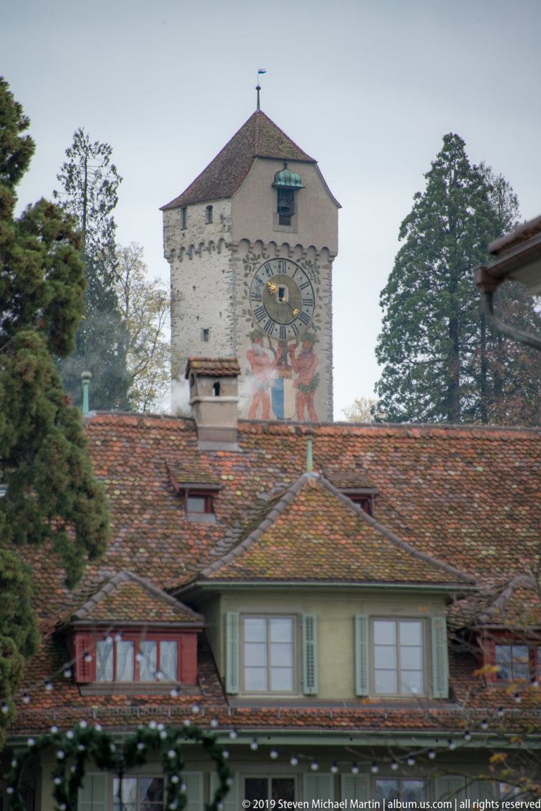 Musegg Wall clock tower rising above tiled roofs, with large clock face and painted decoration.