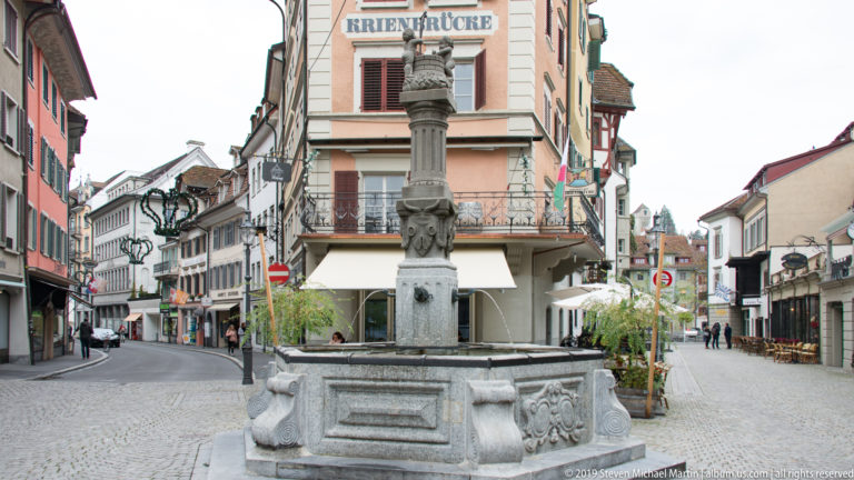 Historic stone fountain with tall sculpted column standing at a Lucerne old-town street junction.