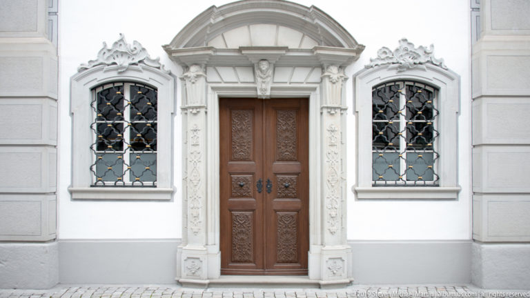 Ornate carved stone doorway with wooden double doors and iron-grilled windows on a white façade.