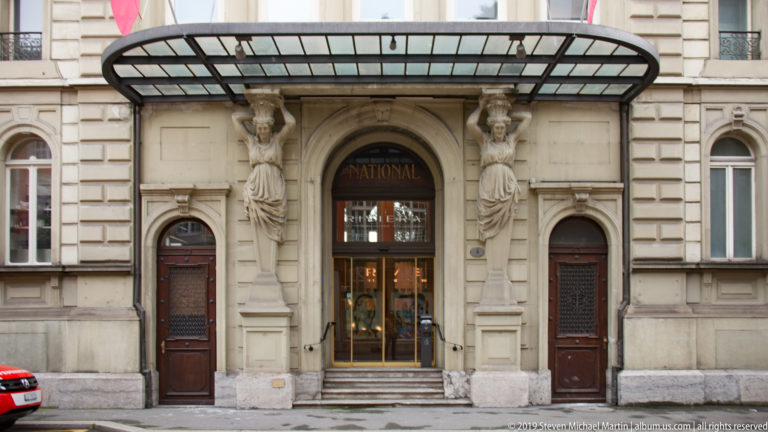 Ornate Grand Hotel National Lucerne entrance with sculpted female figures, canopy, and the word NATIONAL above brass doors.