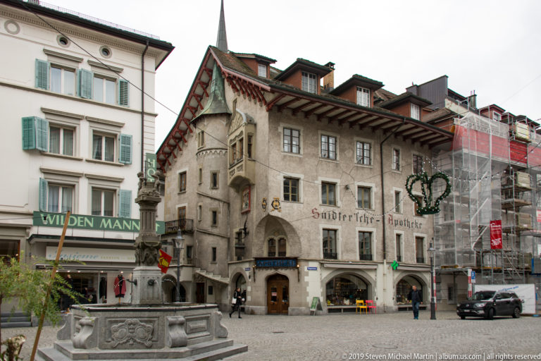 Stone fountain before a historic pharmacy building with projecting eaves in Lucerne’s old town.