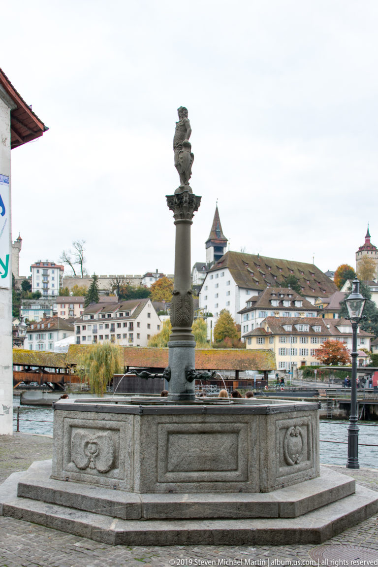 Polygonal stone fountain with central column beside the Reuss, with Lucerne buildings and a hill tower beyond.