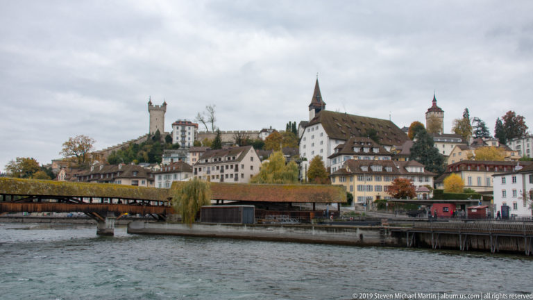 Covered timber bridge along the Reuss with historic hillside buildings, church and defensive towers beyond.