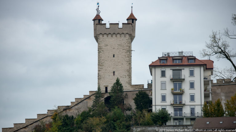 Square stone tower of Lucerne’s Musegg Wall above crenellated walls and nearby residential buildings.