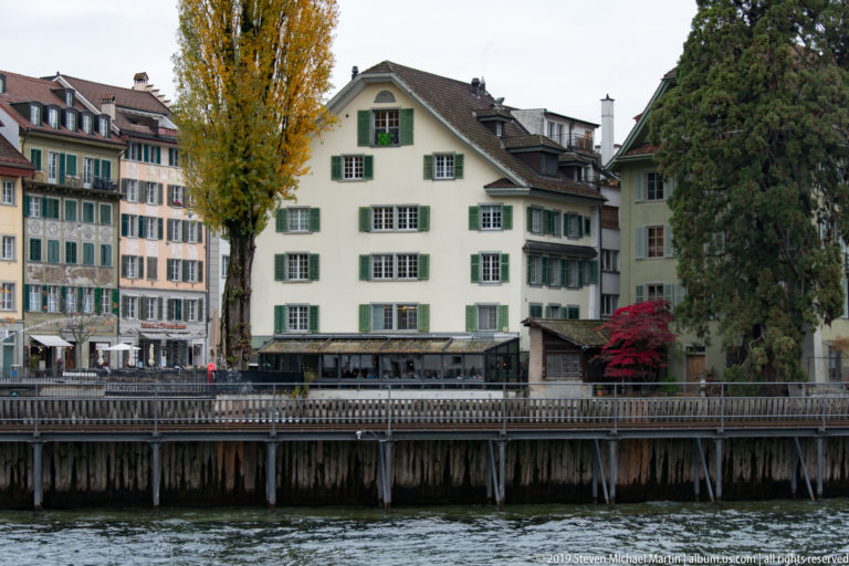 Historic Lucerne houses with shutters and steep roofs lining a walkway beside the Reuss River.