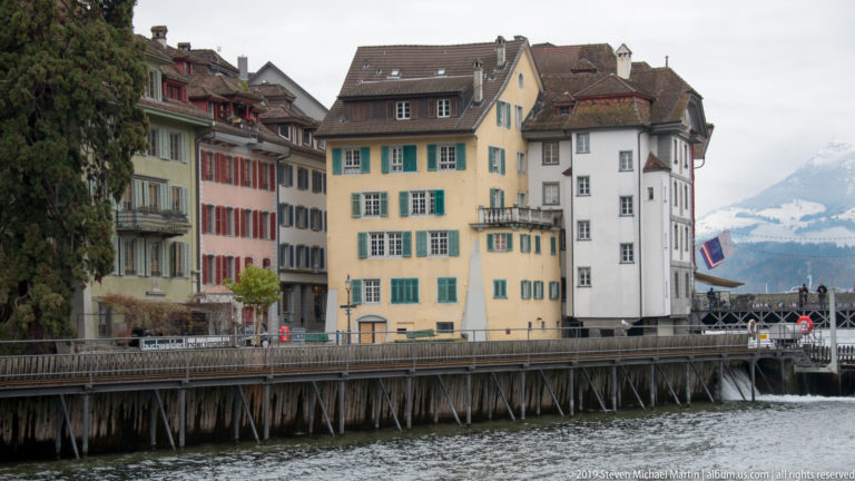 Cluster of tall historic Lucerne houses with colored shutters along the Reuss, with mountains faintly beyond.