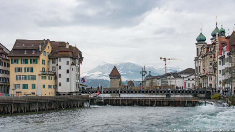 Reuss River with pedestrian bridge, historic buildings, Jesuit Church twin towers, and mountains beyond.