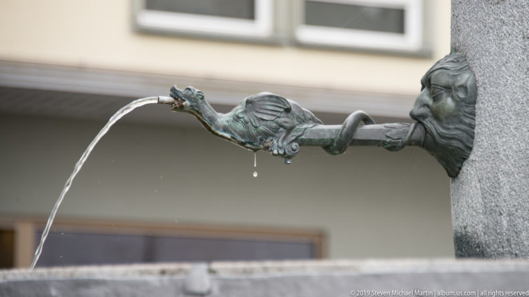 Decorative bronze creature-shaped fountain spout sending an arc of water from a stone basin.