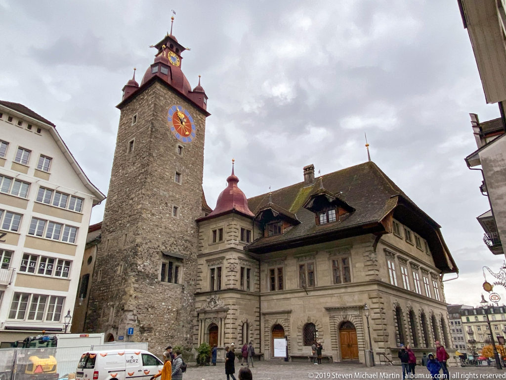 Stone clock tower and adjoining historic Rathaus building in Lucerne, viewed from street level beneath a cloudy sky.