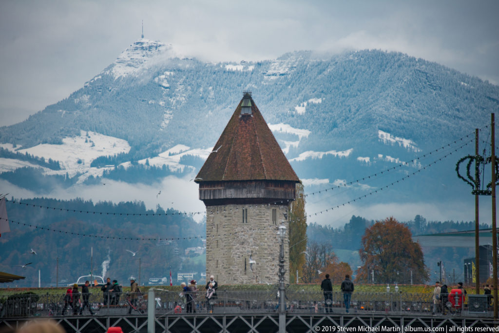 Square stone defensive tower with steep roof beside a bridge, with snow-dusted mountains behind.
