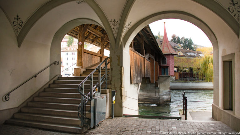 Arched riverside passage and stairway beside Lucerne’s Spreuer Bridge, with timber bridge structure and river beyond.
