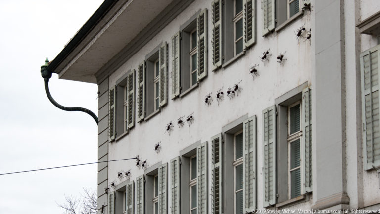 White Lucerne façade with pale shutters, rows of small dark wall ornaments, and curved metal downspout.