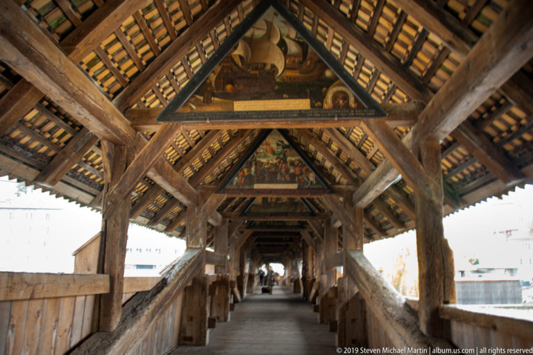 Interior of Lucerne’s Spreuer Bridge with heavy timber framing, covered walkway, and painted triangular roof panels.