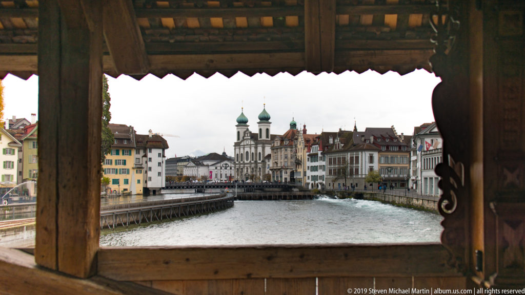 Jesuit Church twin towers and Reuss River framed by the dark timber opening of the Spreuer Bridge.