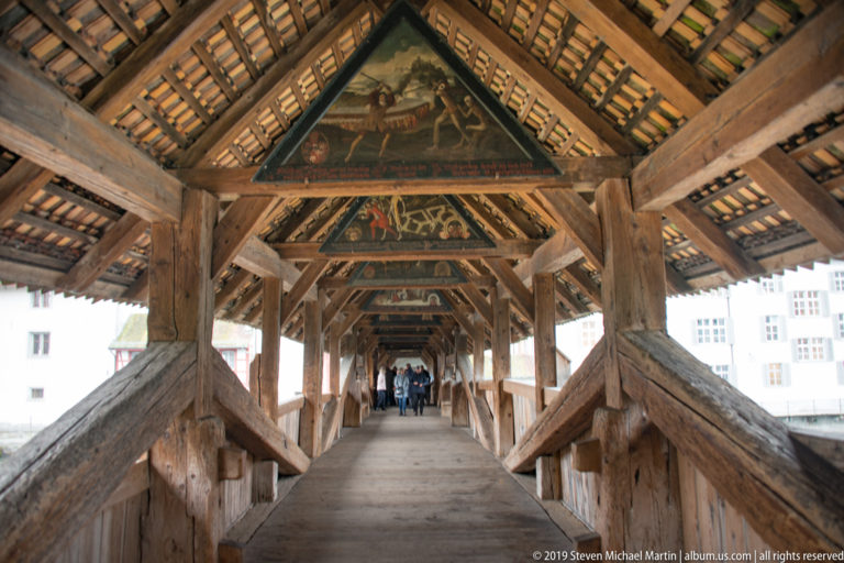 Long interior view through the Spreuer Bridge with painted roof panels and pedestrians in the distance.