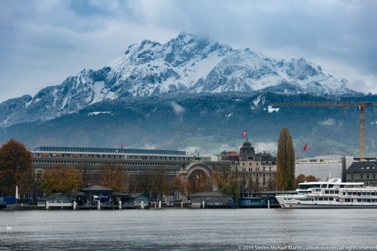 Snow-covered Mount Pilatus above Lucerne waterfront buildings and passenger boats across Lake Lucerne.