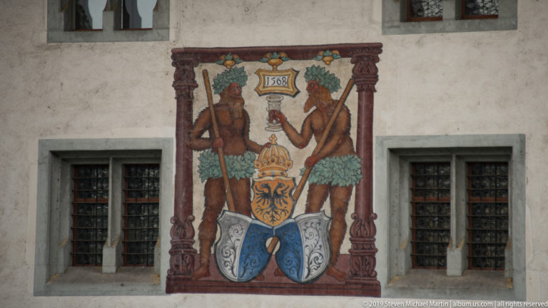 Painted façade mural dated 1568 with two wild men flanking a crowned coat of arms between barred windows.