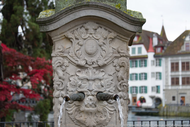 Ornately carved stone fountain with twin metal water spouts, scrollwork relief, and blurred historic buildings behind.