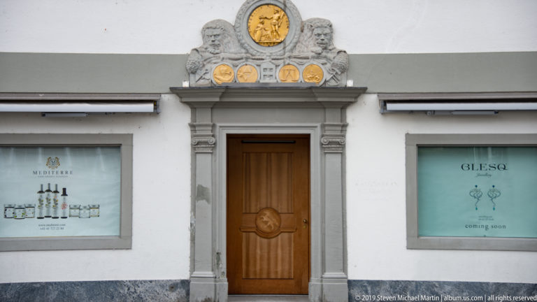 Wooden doorway in a white Lucerne façade, framed by carved stone and topped with gilded heraldic decoration.