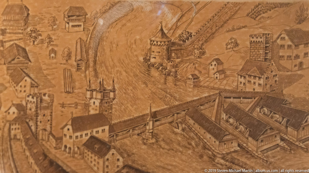 Sepia bird's-eye historical illustration of Lucerne with towers, walls, bridges, and buildings beside the river.