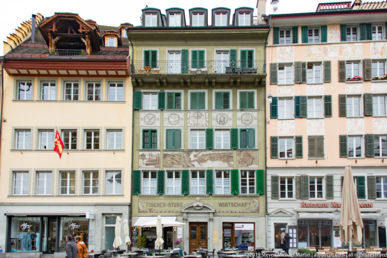 Three adjoining Lucerne buildings with shutters, painted façade decoration, and restaurants at street level.