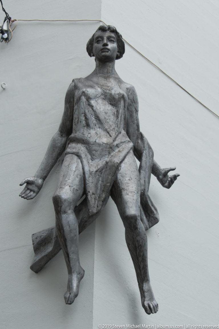Dark metal sculpture of a seated human figure projecting outward from a white wall.