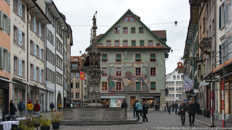 Lucerne old-town square with a sculpted fountain before a green-gabled building covered in painted decoration.