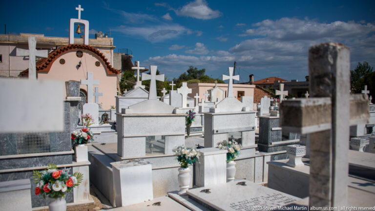 Rows of white and gray cemetery tombs with crosses and flower vases, with a small chapel in the background.
