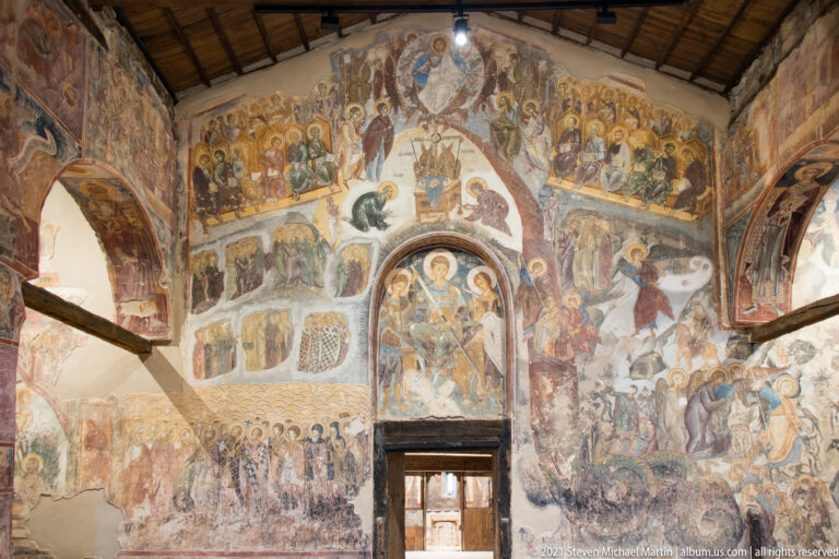 Fresco-covered church wall with multiple saint and narrative scenes surrounding a central doorway beneath a timber roof.