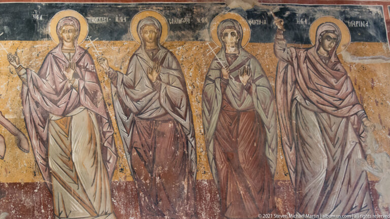 Three standing haloed female saints in long robes and veils against a worn ochre fresco background.