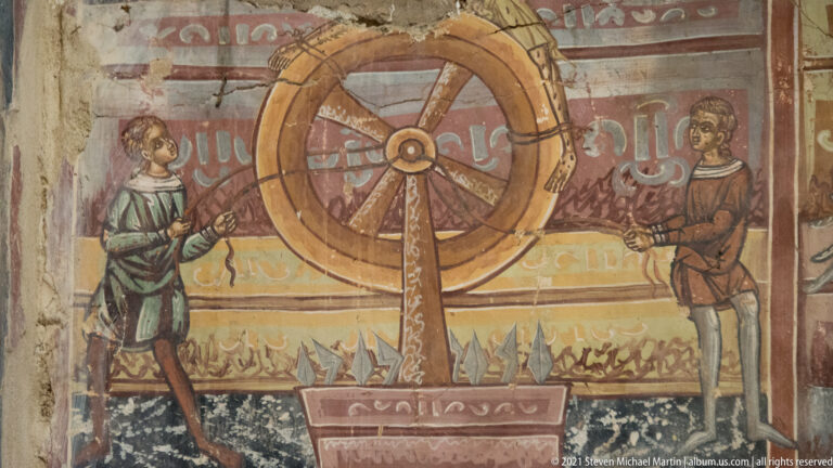 Large wheel on a central support flanked by two attendants, with pointed or flame-like forms along the base.