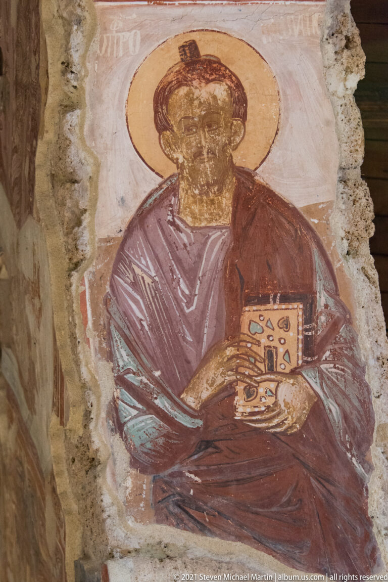 Frontal haloed saint in red-purple robes holding a decorated book against a pale fresco ground.