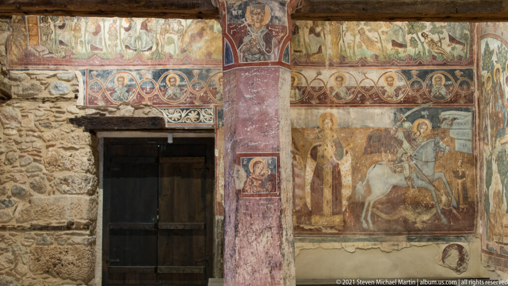 Wooden doorway surrounded by frescoes, including saint medallions above and a mounted warrior scene to the right.