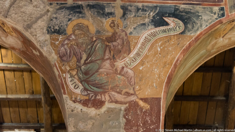 Seated haloed bearded figure on an arch holding a long inscribed scroll across an ochre background.