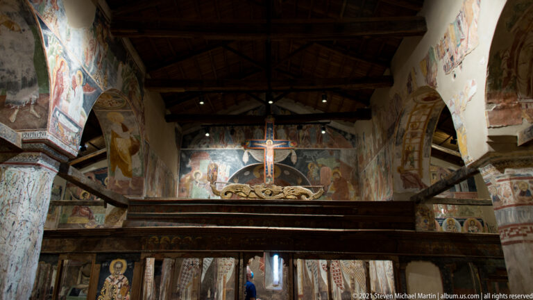 Interior view toward the sanctuary with frescoed walls, timber roof, wooden screen, and a large crucifix on the central axis.