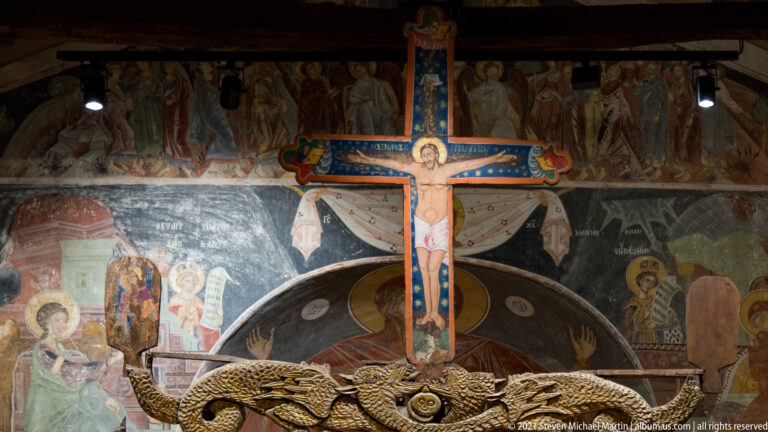 Large brightly painted crucifix before dark sanctuary frescoes, with an ornate wooden liturgical element across the foreground.
