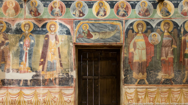 Wooden doorway flanked by full-length saints, with a Jonah sea-creature fresco above and saint medallions across the top.