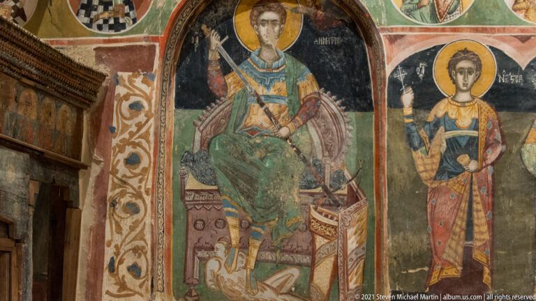 Enthroned haloed warrior saint in elaborate dress beside a standing young haloed saint raising one hand.