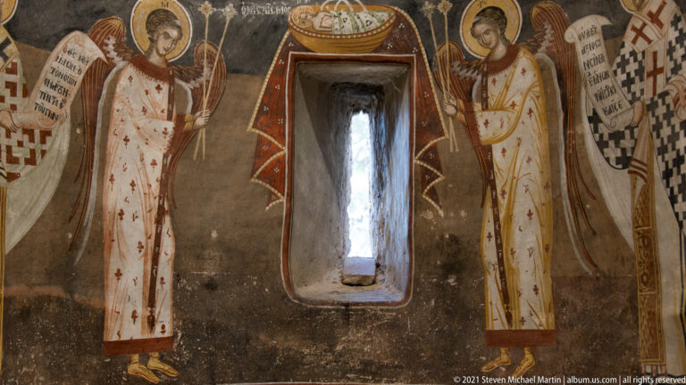 Narrow recessed window between painted standing saints and bishops in patterned vestments on a dark interior wall.