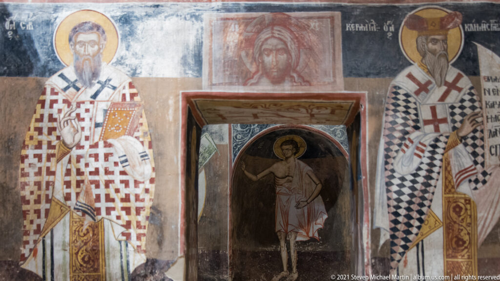 Arched doorway framed by painted bishops, with another standing haloed saint visible in the dark opening.