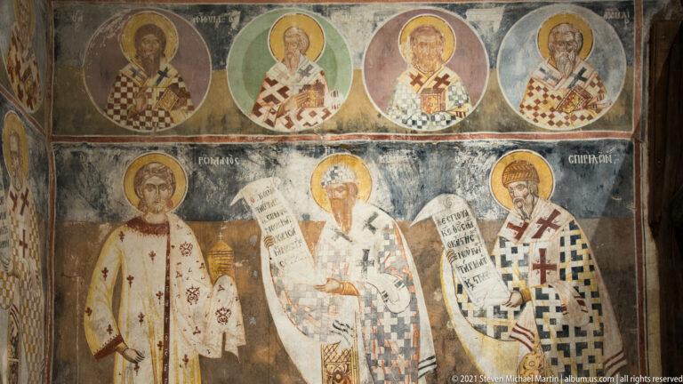 Two mural registers of haloed bishops and saints in patterned vestments, several holding inscribed scrolls.