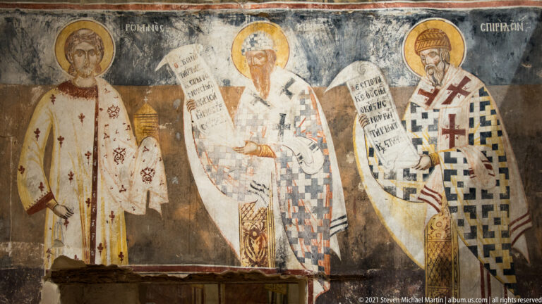Three haloed bishops in patterned vestments, two holding inscribed scrolls, on a dark frescoed wall.