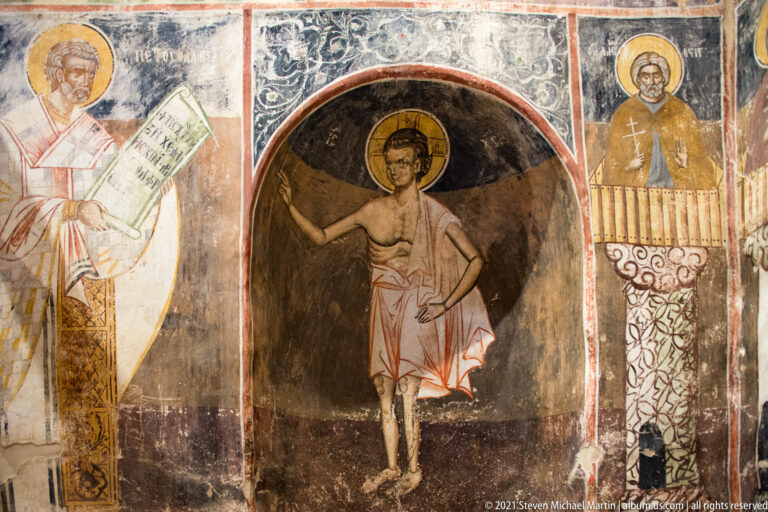 Young haloed saint standing in a dark arched niche, framed by adjacent frescoes and painted ornament.