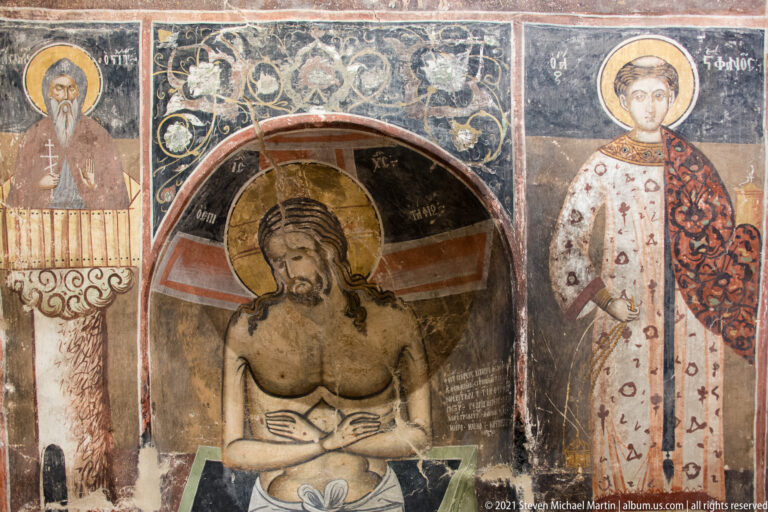 Half-length Christ with bowed head and crossed arms in a dark arched niche beside a standing saint.