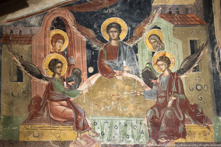 Two winged angels flank a haloed youthful figure before stylized painted buildings and additional saints.