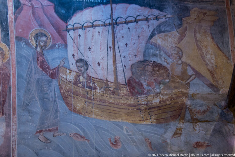 Frescoed sailboat carrying several figures beneath a large pale sail, with a rocky form at right.