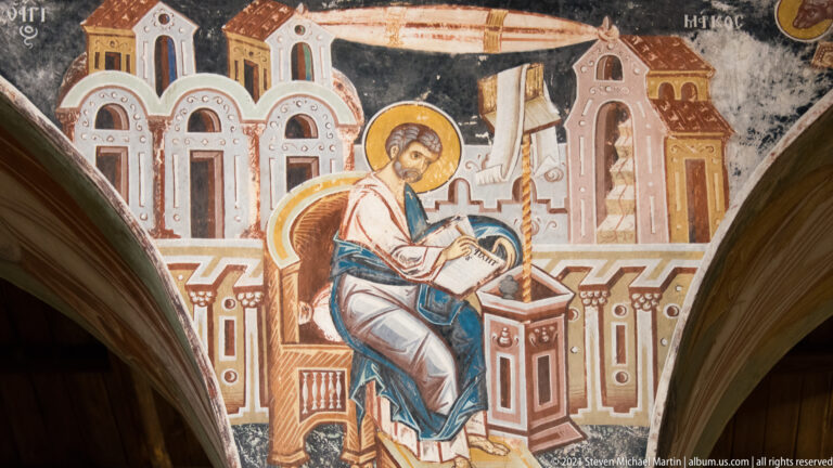 Seated haloed evangelist writing in an open book at a lectern before stylized painted architecture.