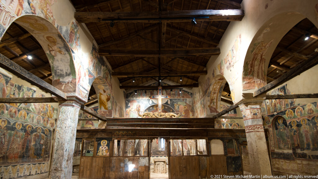Interior of the church with frescoed arcades, painted columns, timber roof, and a wooden screen across the nave.